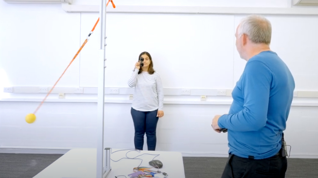 Person holding sunglasses over one eye whilst watching a ball swing forwards and backwards on a string.